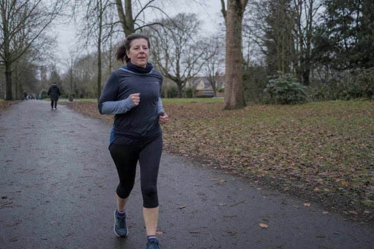 A woman in her mid-fifties runs at an easy pace along a park path on an overcast morning, wearing casual layers and trainers, clearly not racing