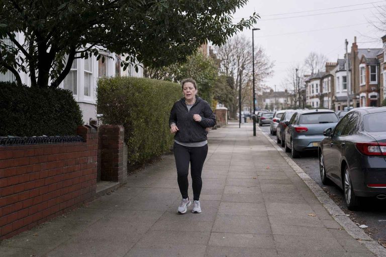 A beginner runner on a wide suburban pavement on a grey UK morning, viewed from the side, wearing ordinary everyday kit