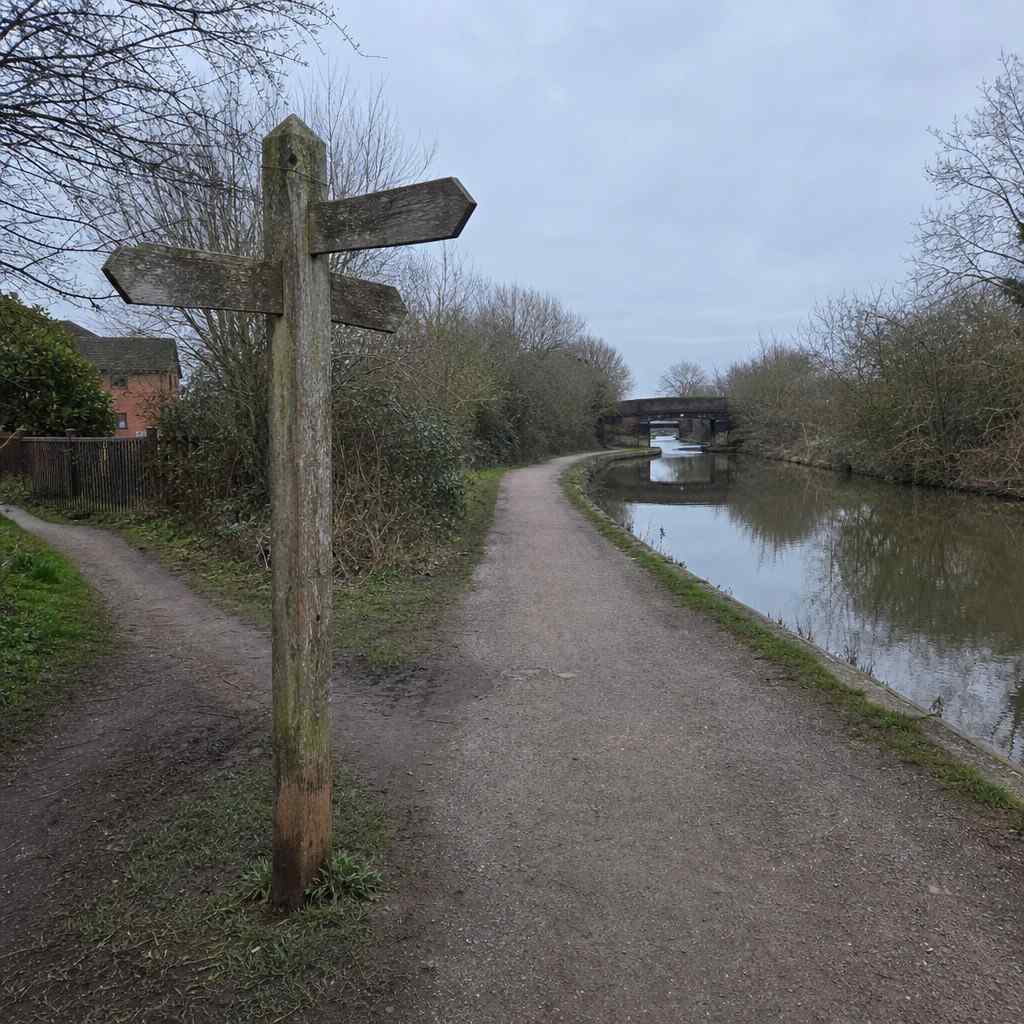 A canal towpath junction in the UK with a wooden signpost, flat path ahead, grey morning light, no runners in shot