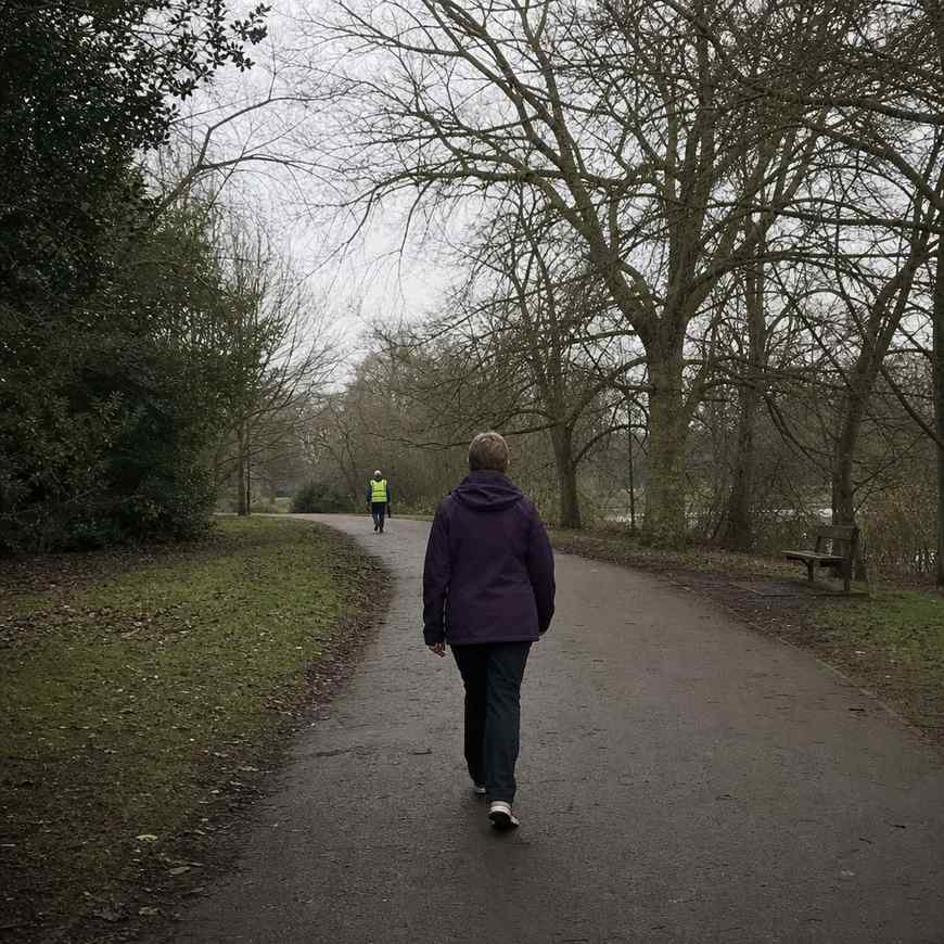 A lone walker near the back of a parkrun course on a grey morning in a UK park, with a volunteer tail walker behind them