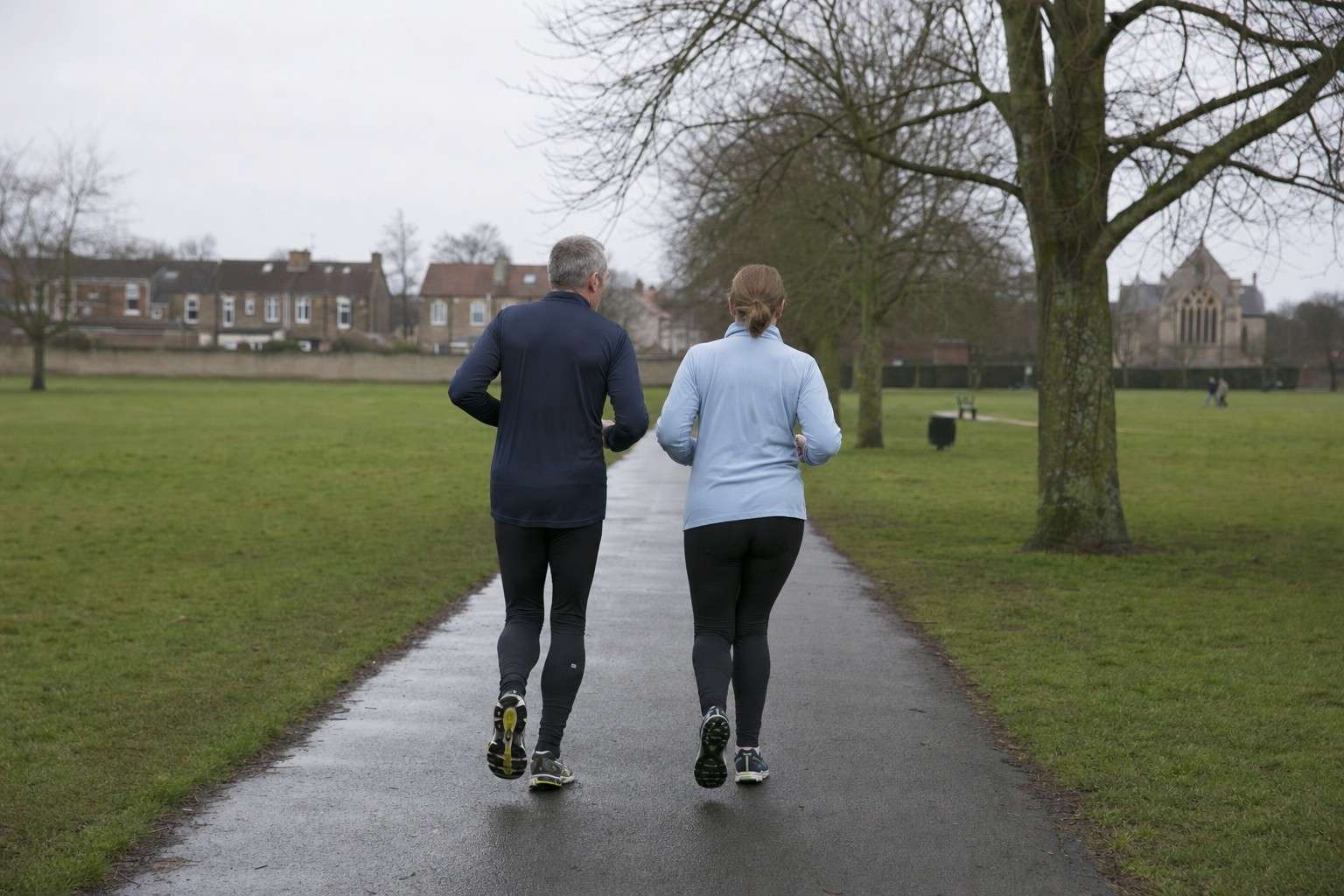 Two ordinary runners in their fifties jogging slowly side by side along a park path on a grey morning, seen from behind