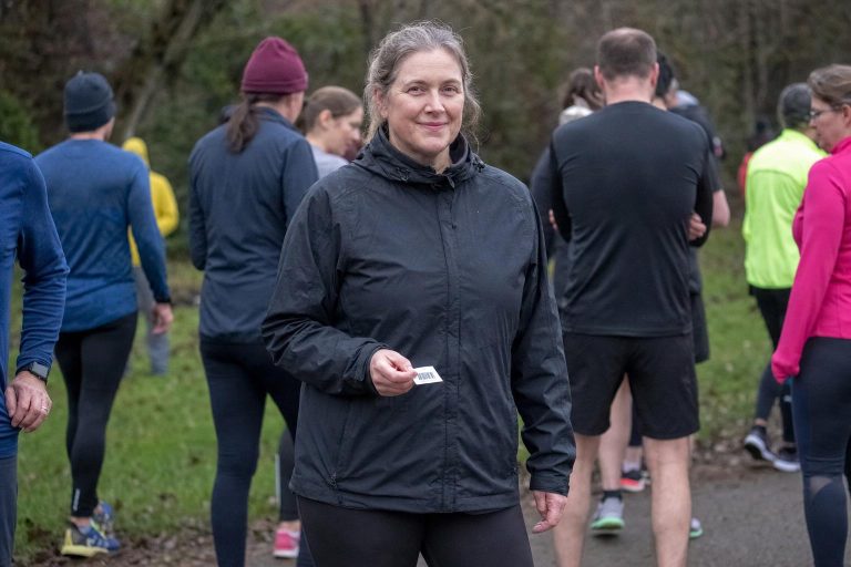 A woman in her early 50s at a parkrun start line on a grey Saturday morning, holding a barcode printout