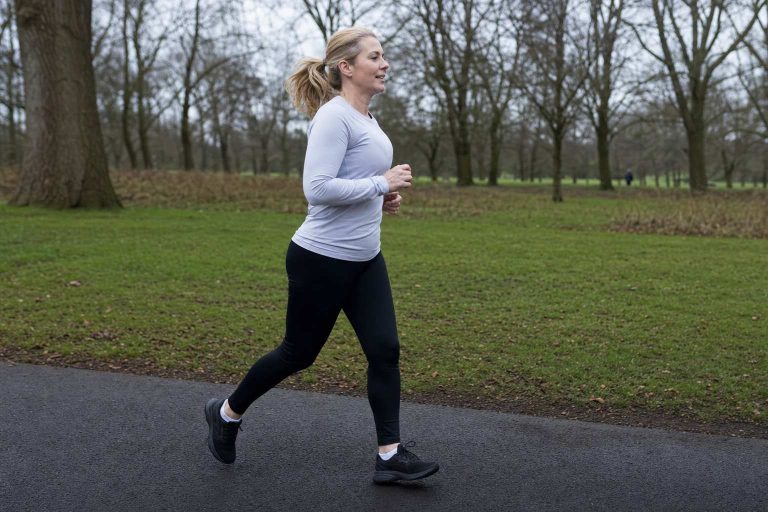 A woman in her 50s running at an easy, relaxed pace along a park path, clearly comfortable and unhurried