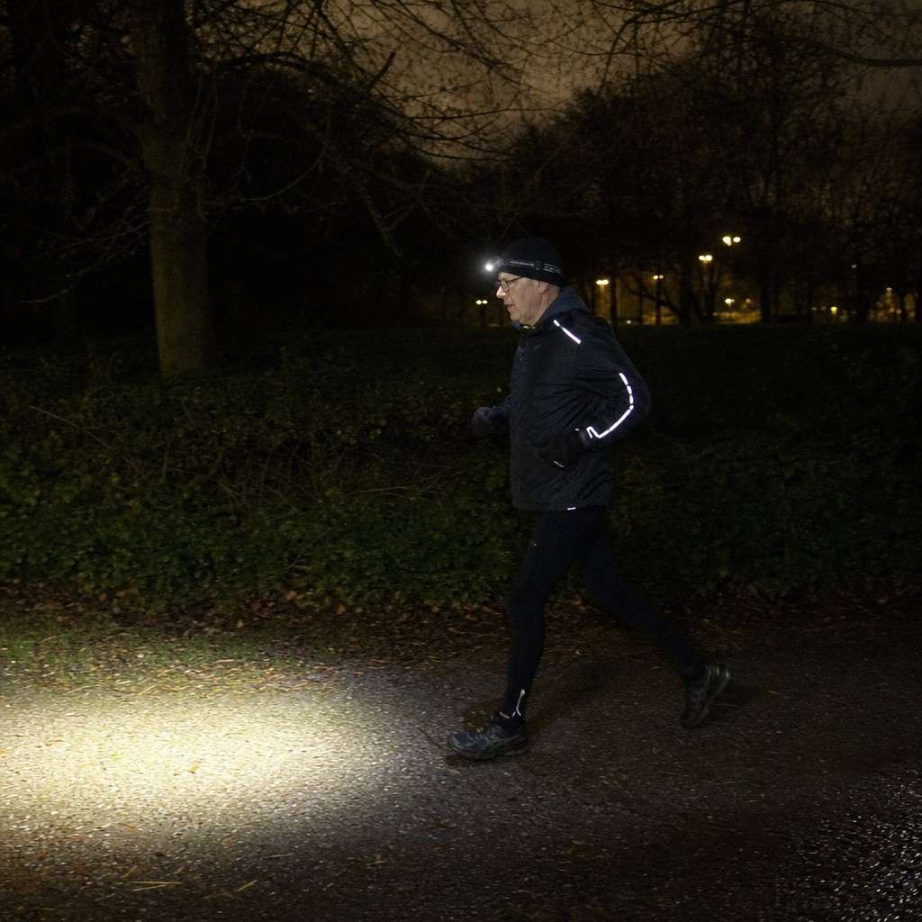 Runner with a head torch on an unlit park path at night, beam visible on the ground ahead