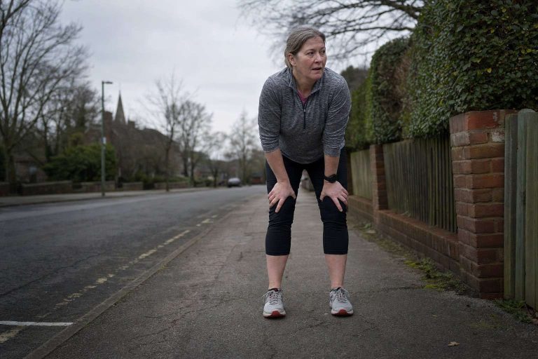 A woman in her fifties pausing during a run on a suburban pavement, hands on her knees, catching her breath on an overcast morning