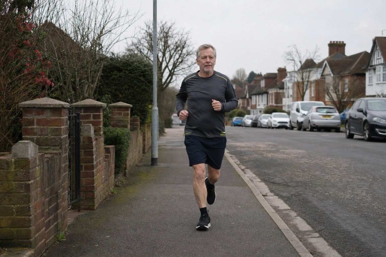 A runner in their late fifties jogging on a grey suburban pavement on an overcast morning